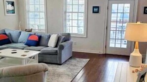 A living room with a gray sofa, a white coffee table, and a patterned rug. There are two windows with blinds, a ceiling fan, and a white side table with a lamp. - Cape Haven - Condo 1.5 Blocks from Beach (Cape Charles)