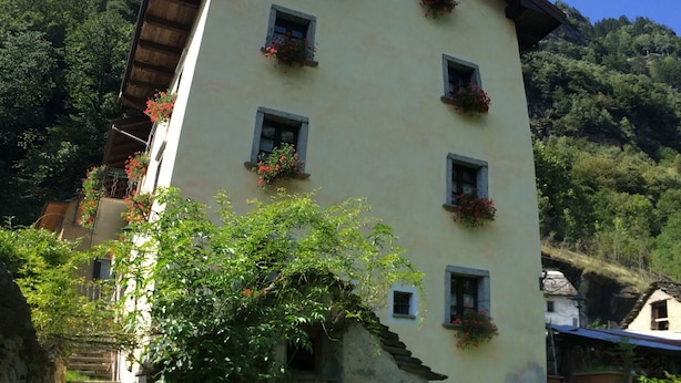 Um edifício com várias janelas, algumas com caixas de flores. Há uma mesa com cadeiras montada do lado de fora.