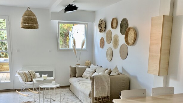 Un salon moderne avec un canapé, une table à manger et une table basse. On y trouve des paniers tressés accrochés au mur et une fenêtre donnant sur un paysage verdoyant.