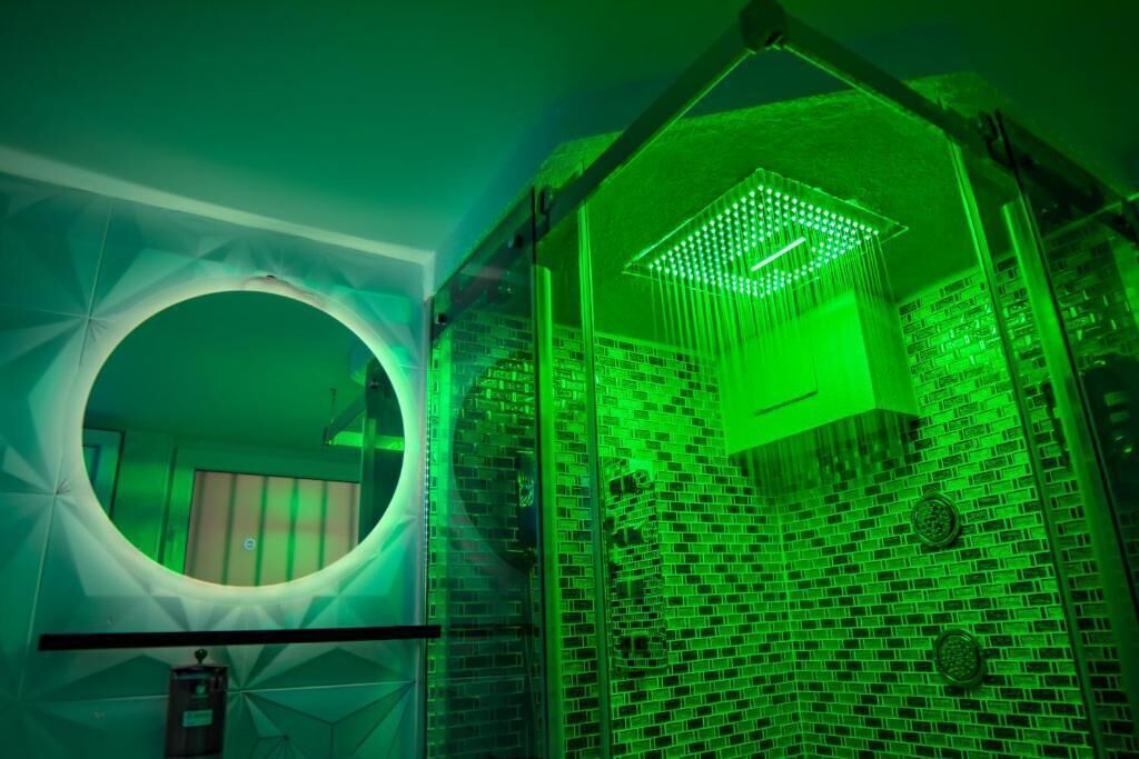 A room with a circular mirror, a green-lit shower area, and a brick-patterned wall.