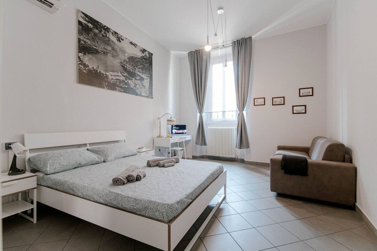 A modern bedroom with a bed, a desk, a chair, a sofa, and a window with curtains.