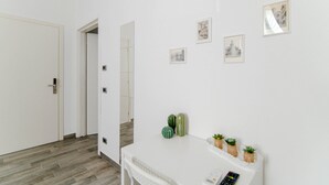 Un pasillo con una puerta blanca, una pared blanca decorada con marcos y una mesita con plantas y un control remoto.