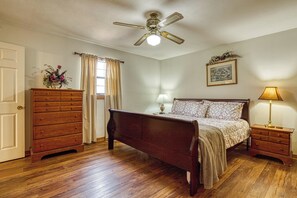 Un dormitorio con cama de madera, mesita de noche, cómoda, ventilador de techo, ventana con cortinas e imagen enmarcada en la pared.