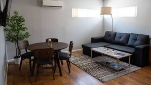 Ein Wohnzimmer mit einem runden Esstisch, Stühlen, einem schwarzen Ledersofa, einem Couchtisch, einer Stehlampe und einem Blumentopf.