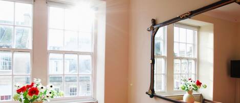 A room with a large window, a mirror, and two vases with flowers.