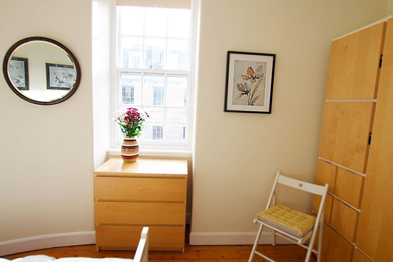 A bedroom with a window, a dresser, a chair, a mirror, and a framed picture on the wall.