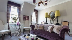 A living room with a purple sofa, a round coffee table with a tray of tea and biscuits, a white armchair, and a small table with a lamp. - Charming Nest for Two with Castle View (Edinburgh)