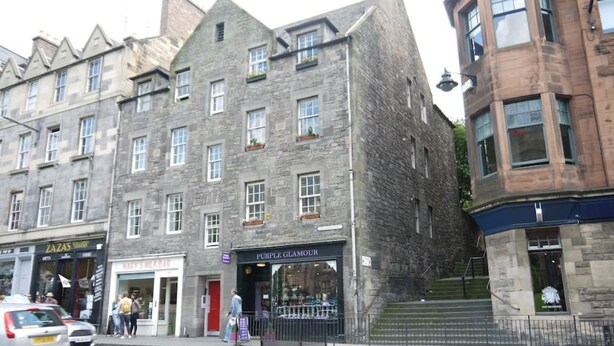 A multi-story stone building with shops on the ground floor, including a jewelry store and a milk bar. - Charming Nest for Two with Castle View (Edinburgh)