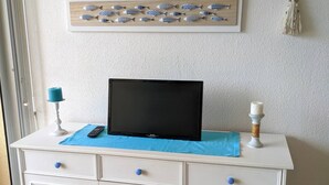 A white cabinet with a flat-screen TV on top, a decorative fish wall hanging, and a small table with a blue cloth. - Large sea-facing studio with balcony and outdoor parkingeur 4 people (Saint-Jean-de-Monts)