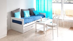 A bright room with a white bed, a ceiling fan, a window with blue curtains, and a white chair. - Large sea-facing studio with balcony and outdoor parkingeur 4 people (Saint-Jean-de-Monts)