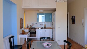 A compact kitchen with white cabinets, a stove, and a sink. There is a dining table with chairs and a bowl of fruit. - Casa Naumachie Relax in a Safe and Wonderful Place (Taormina)