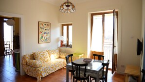 A living room with a sofa, dining table, chairs, a painting, and a balcony door. - Casa Naumachie Relax in a Safe and Wonderful Place (Taormina)