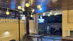 Un área de comedor al aire libre con piso estampado, mesas y sillas de madera, luces esféricas colgantes y la entrada visible a un restaurante.
