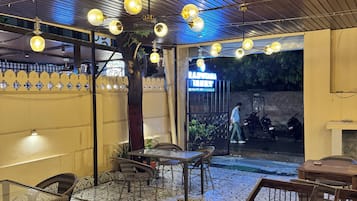 Uma área de refeições ao ar livre com piso estampado, mesas e cadeiras de madeira, luminárias esféricas pendentes e entrada visível para um restaurante.