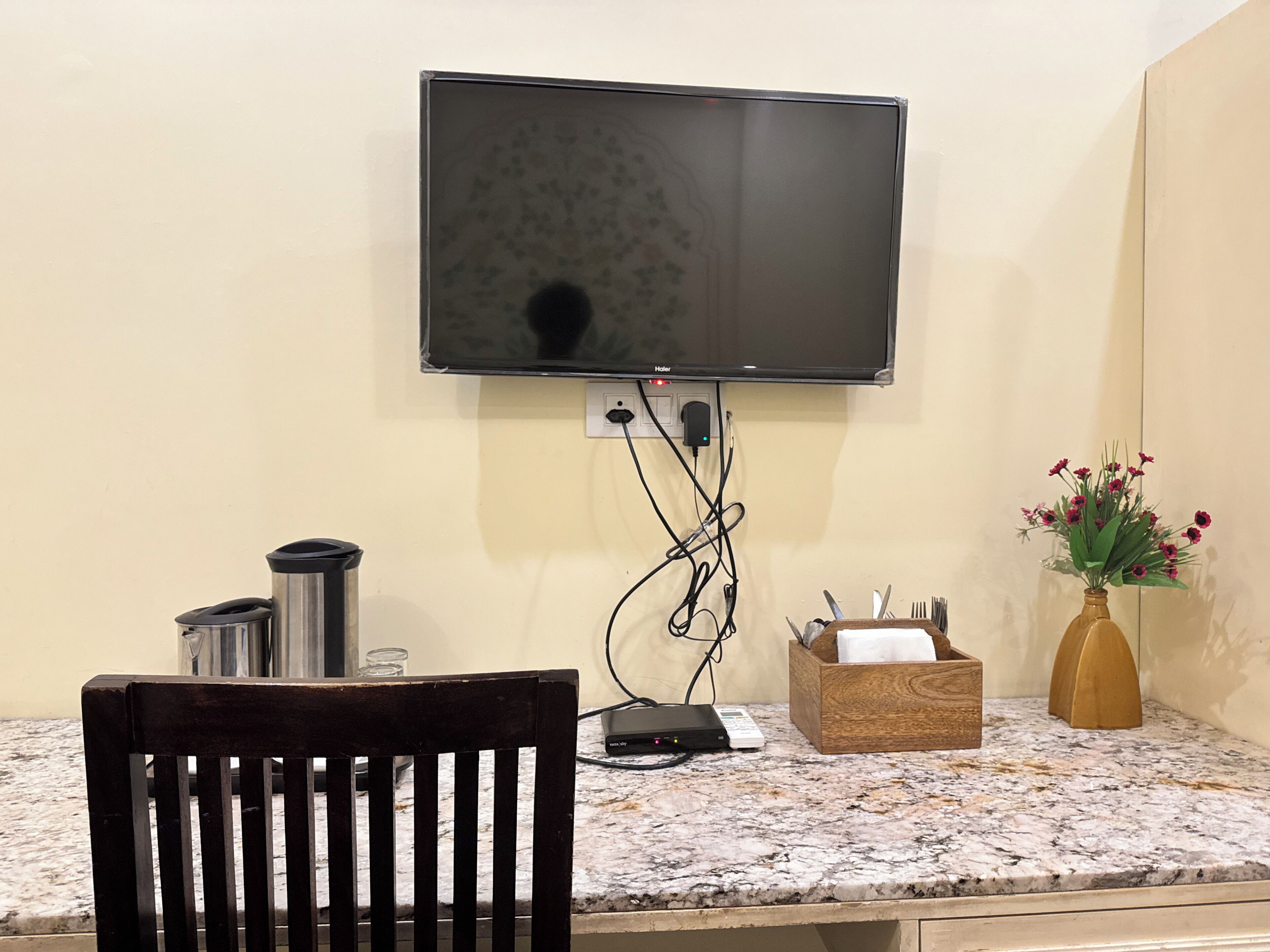 Televisão de tela plana na parede, uma cadeira de madeira, bancada de mármore, um vaso com flores e uma caixa com utensílios.