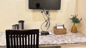 A flat-screen TV mounted on a wall, a wooden chair, a marble countertop, a vase with flowers, and a box with utensils. - Rajputana Diaries (Jaipur)