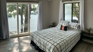 Ein Schlafzimmer mit einem großen Bett, zwei Nachttischen, einem Fenster mit Blick auf Bäume und Wasser sowie einer Zimmerpflanze auf einem Tisch.