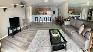 Ein modernes Wohnzimmer mit einem grauen Sofa, einem schwarzen Couchtisch mit einer Pflanze, einem Fernseher auf einem Schrank und einer Küche im Hintergrund.