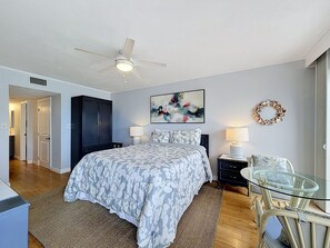 Ein Schlafzimmer mit einem Bett, zwei Nachttischlampen, einem Deckenventilator, einem Glastisch, einem Sessel, einem Wandbild und einem dekorativen Kranz.