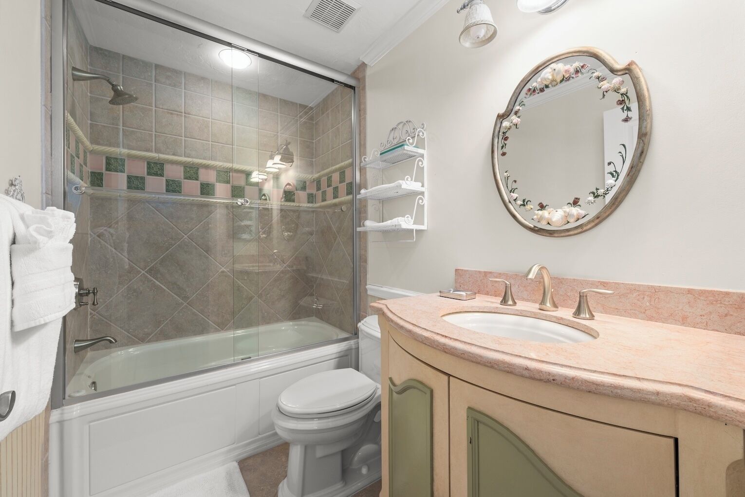 Een badkamer met een douchecabine van glas, een wit toilet, een wastafel met roze aanrechtblad en een spiegel met bloemenmotief.