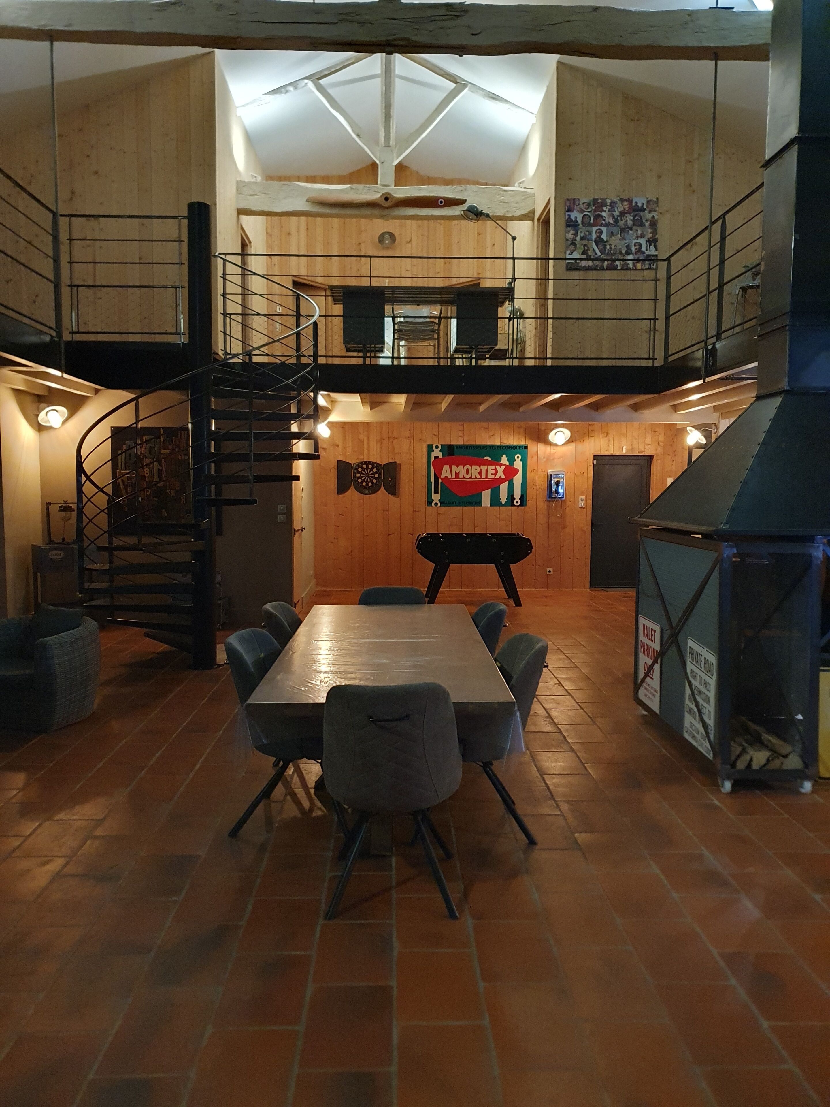 Un vaste espace dédié à la salle à manger, doté d’un haut plafond et de murs en bois, comprenant une table et des chaises, un escalier et une enseigne indiquant « AMORTEX ».