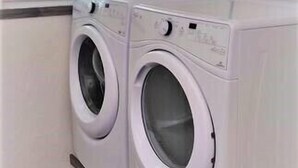 A laundry room with two white front-loading washing machines, a window with blinds, and a power outlet on the wall.
