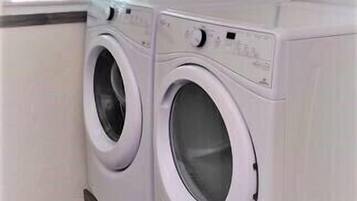 Une buanderie équipée de deux machines à laver à chargement frontal blanches, d’une fenêtre avec des stores et d’une prise électrique au mur.
