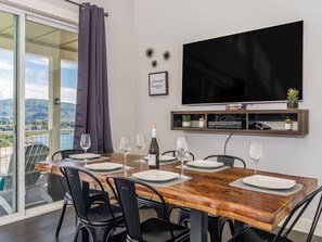 Ein Essbereich mit einem Holztisch, gedeckt für vier Personen, schwarzen Stühlen, einem an der Wand befestigten Fernseher und einem schönen Ausblick durch die Glasdoor.