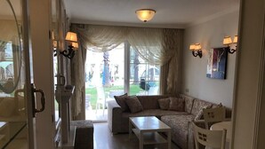 Ein Wohnzimmer mit einem Ecksofa, einem kleinen Tisch und einem Sessel. Eine Glastür führt zu einem Außenbereich mit Rasen und Sitzgelegenheiten.