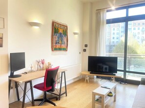 A modern living room with a large flat-screen TV, a desk with a computer, a red chair, and a window with a view of buildings.