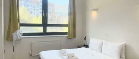 A hotel room with a bed, a window with curtains, a radiator, a standing fan, and a nightstand with a lamp.