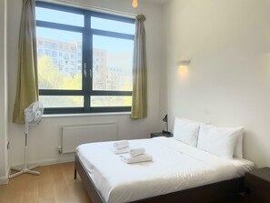 A hotel room with a bed, a window with curtains, a radiator, a standing fan, and a nightstand with a lamp.