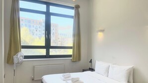 A hotel room with a bed, a window with curtains, a standing fan, a bedside lamp, and a view of buildings outside. - Chic 1 BDR APT w Balcony in Shepherd's Bush (London)