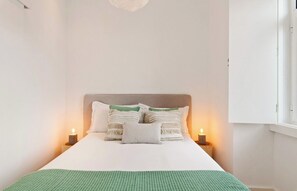 A neatly made bed with a white sheet and a green blanket, flanked by two bedside tables with candles, in a room with white walls and a window. - Modern 1BDR Stay in Charming Belém (Lisboa)
