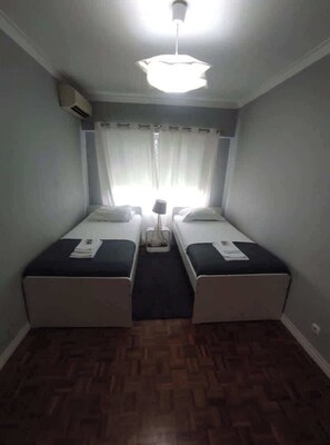 A room with two single beds, a ceiling fan, an air conditioning unit, and a window with curtains.