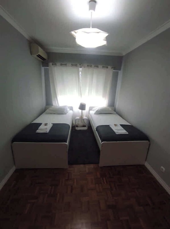 A room with two single beds, a ceiling fan, an air conditioning unit, and a window with curtains.