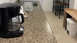 A compact kitchen with a granite countertop, a coffee maker, a refrigerator, and a small dining area with a table and chairs. - Be At Home Away From Home III (Welland)