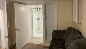 A room with a closed door, a glass shower door, a beige wall, and a dark-colored sofa. - Be At Home Away From Home III (Welland)