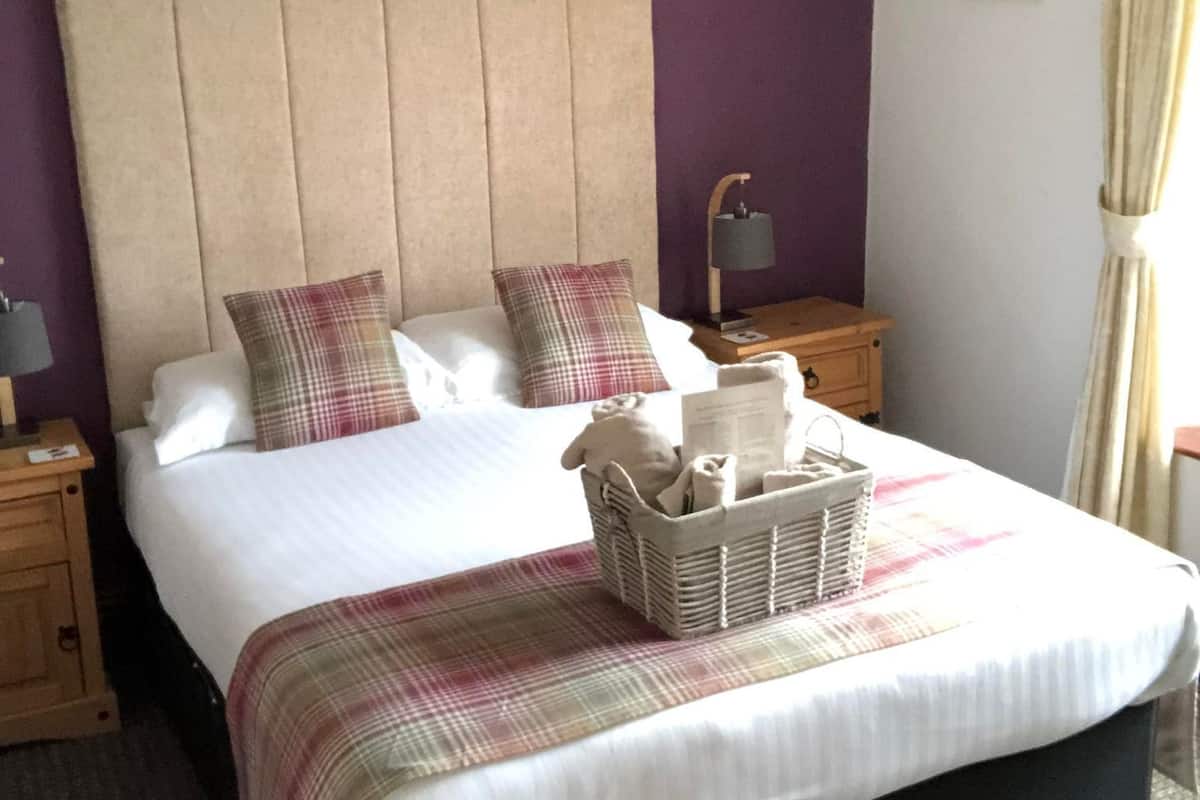 Un lit bien fait, recouvert d’une couverture à carreaux, une lampe de chevet et un panier contenant des articles de toilette sur la table de chevet.