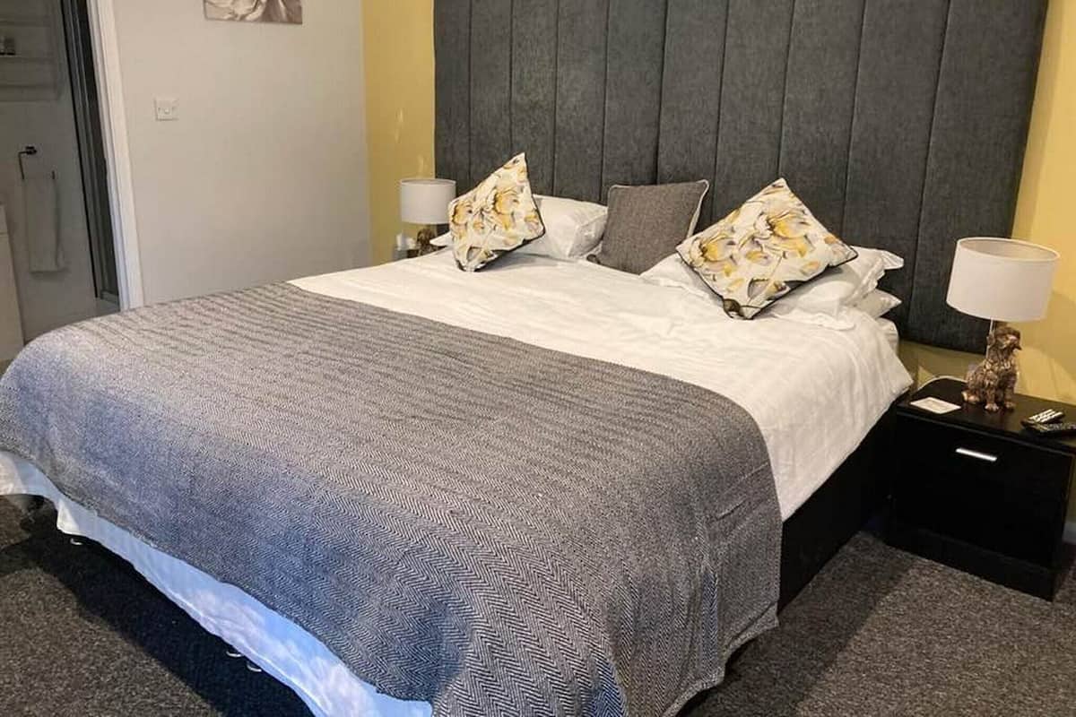 Un lit bien fait, recouvert d’une couvre-lit à motifs gris et blancs, avec deux oreillers et une tête de lit grise. On y trouve deux tables de chevet avec des lampes, et un petit objet décoratif sur l’une d’elles.