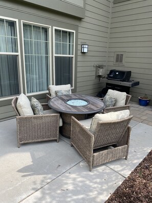 Relax outdoors with a round table and four chairs on this patio, adjacent to a building with large windows and a grill.