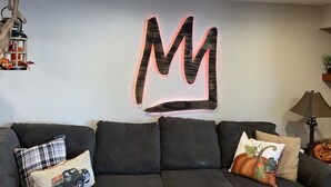 A living room with a dark grey sofa, a large illuminated 'M' wall art, and a hanging lantern with autumn leaves.