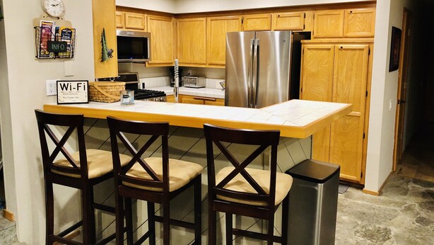 A kitchen with wooden cabinets, a stainless steel refrigerator, a microwave, and a sink. There are three bar stools and a Wi-Fi sign on the counter.