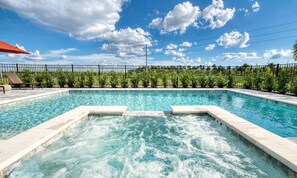 A swimming pool with a hot tub in the center, surrounded by a fence and greenery.