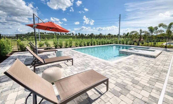 A swimming pool with lounge chairs and a red umbrella, surrounded by a paved patio and a fence.