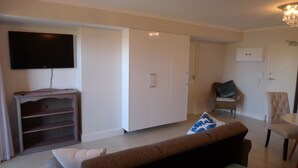 A living room with a flat-screen TV, a white wardrobe, a wicker chair, a brown sofa, and a small table. - Neat Modern Studio in Safe Complex in the Heart of Claremont (Cape Town)