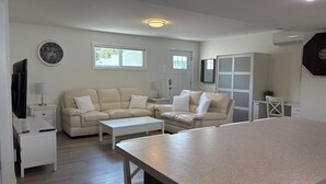 Un salon moderne avec un canapé beige, une table basse et un téléviseur posé sur un meuble. On y trouve une fenêtre qui laisse entrer la lumière naturelle, une porte et une petite plante sur un bureau.