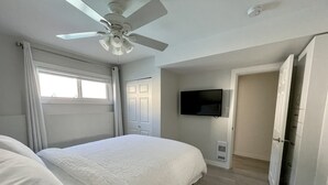 Une chambre à coucher comprenant un lit, un ventilateur de plafond, une télévision et une porte donnant sur une autre pièce.