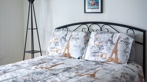 A bed with a Paris-themed duvet and pillows, a black metal headboard, a framed picture on the wall, and a tall floor lamp.