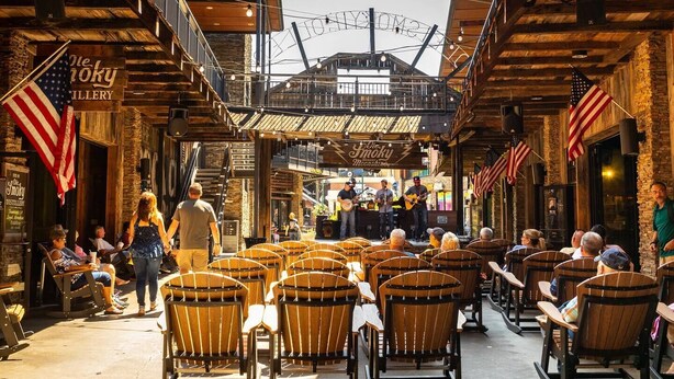 An outdoor seating area with wooden chairs and tables, a stage with musicians, and American flags.